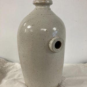 Puede incluir: Una olla de cerámica blanca con una abertura redonda en el lateral. La olla tiene un esmalte moteado y un pequeño agujero en el lateral. La olla está sentada sobre un mantel de encaje blanco.