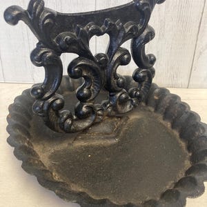 May include: A black, cast iron decorative dish with a scalloped edge and ornate, scrollwork design. The dish has a heart-shaped base and a curved, openwork handle. The finish is matte and slightly distressed, giving it a vintage look.