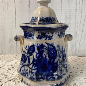 May include: A vintage ceramic jar with a lid, featuring a blue and white floral pattern. The jar has a bulbous shape with two small handles and a decorative finial on the lid. The overall design is reminiscent of antique pottery.