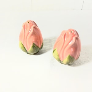 May include: A pair of ceramic salt and pepper shakers shaped like closed rosebuds. Each shaker features a glossy, light pink petal exterior with a green, leaf-like base. The top of each rosebud has a small hole for dispensing seasoning.