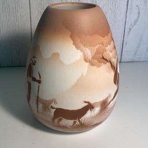 Może przedstawiać: Matowy, jajowaty wazon ceramiczny z jasnobrązowym i białym gradientem w tle. Powierzchnia ozdobiona jest ciemnobrązową ilustracją sylwetkową przedstawiającą osobę trzymającą laskę oraz dwie kozy w krajobrazie.