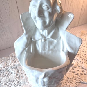 May include: A white ceramic wall pocket featuring a sculpted bust of a historical figure with a high-collared coat. The piece has a hollow, open-top compartment integrated into the front, designed for holding small items or floral arrangements.