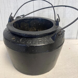 Könnte beinhalten: Ein schwarzer gusseiserner Kessel mit rundem Körper und kreisförmiger Öffnung. Der Kessel hat einen Drahtgriff und wird wahrscheinlich zum Kochen oder zu dekorativen Zwecken verwendet. Das Wort "COPA" ist auf der Seite geprägt.