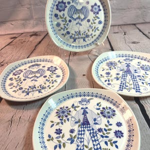 May include: A set of four white ceramic plates featuring blue and green line art illustrations. Two plates depict a figure in a dress, and two plates depict a figure in a striped shirt and checkered pants, all surrounded by floral motifs.
