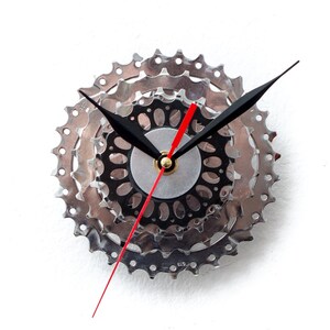 May include: A unique wall clock crafted from recycled bicycle gears. The clock features a silver gear design with black and red clock hands. The clock hands are black, and the second hand is red. The clock is mounted on a white background.