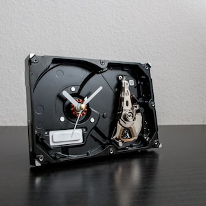 Könnte beinhalten: Eine schwarze Festplatte, die zu einer Uhr mit weißen Zeigern umgebaut wurde. Die Festplatte ist geöffnet und zeigt die inneren Komponenten.