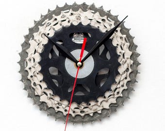May include: A unique wall clock crafted from bicycle gears. The clock features a black center gear, surrounded by silver and gray gears, with black clock hands and a red second hand. The clock is mounted on a white background.