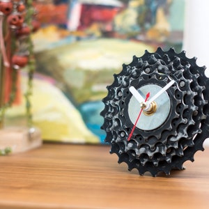 Op de afbeelding: Een zwarte en zilveren klok gemaakt van een fietskettingblad. De klok heeft witte wijzers en een rode secondewijzer.