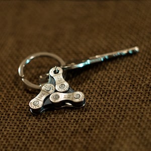 May include: A silver key chain with a triangular bike chain link charm. The chain link has the words "IGSI" and "Shimano" printed on it.