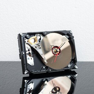 May include: A repurposed computer hard drive converted into a wall clock. The device features a circular silver platter, a metallic actuator arm, and a clock face mechanism with black hour and minute hands and a thin red second hand.