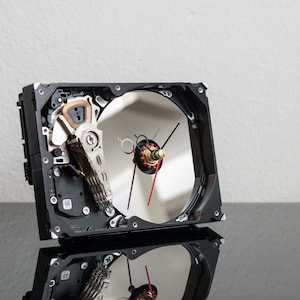 Puede incluir: Un reloj único hecho de un disco duro de computadora reutilizado. El reloj presenta una esfera plateada reflectante, manecillas negras y un segundero rojo. Los componentes mecánicos del reloj son visibles.