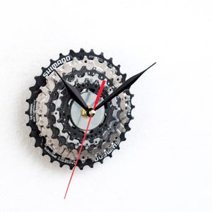 以下が含まれることがあります： 自転車のギアで作られたユニークな壁掛け時計。 黒とシルバーのギアデザインで、黒い針と赤い秒針が付いています。 文字盤にはShimanoのブランド名が表示されています。