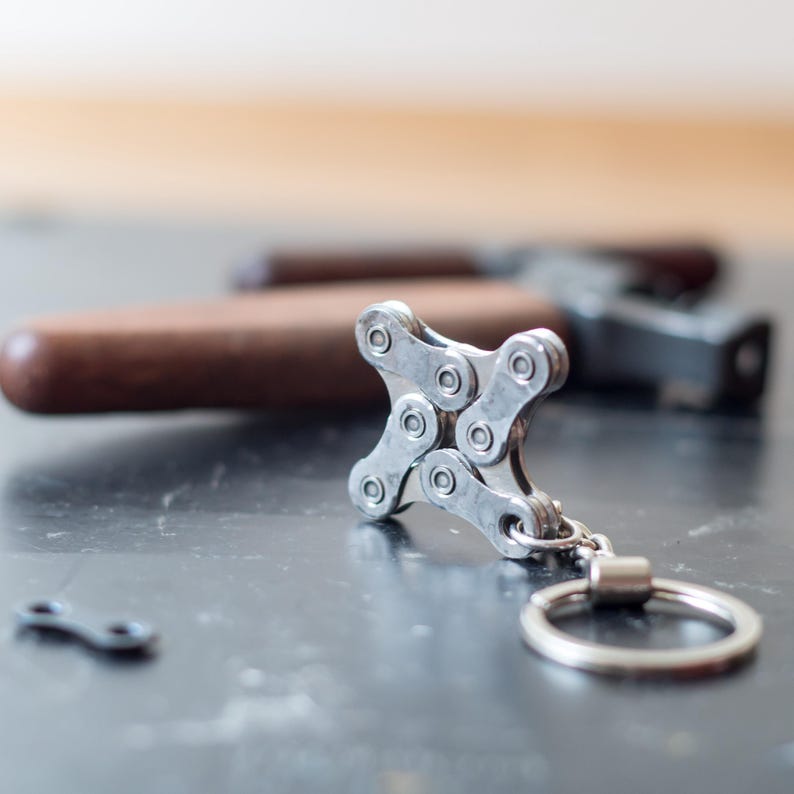 May include: A silver bike chain keychain with a silver ring. The keychain is shaped like a cross.