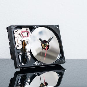 May include: A wall clock constructed from a repurposed hard drive, featuring a black rectangular frame, a circular silver platter, and internal mechanical components. The clock face includes black hour and minute hands with a thin red second hand.