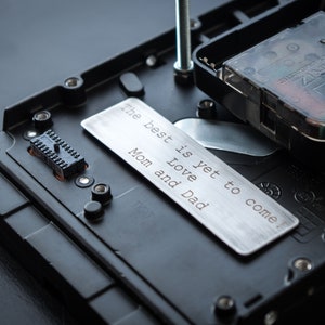 May include: A black quartz clock mechanism mounted on a black plastic base, featuring a rectangular silver metal plate engraved with the text: The best is yet to come! Love Mom and Dad. A vertical metal screw extends upward from the base.