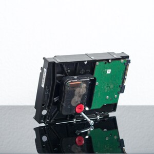 May include: A black computer hard drive repurposed as a clock, featuring a green circuit board on the back, a black plastic clock mechanism mounted in the center, and a small red gear attached to a silver metal clock hand spindle.