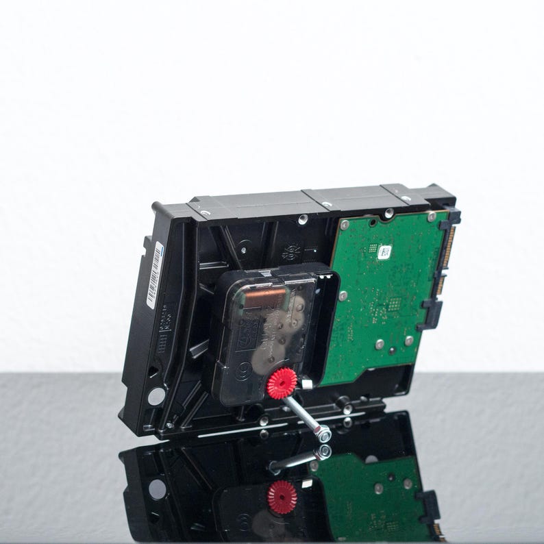 May include: A black computer hard drive repurposed as a clock, featuring a green circuit board on the back, a black plastic clock mechanism mounted in the center, and a small red gear attached to a silver metal clock hand spindle.