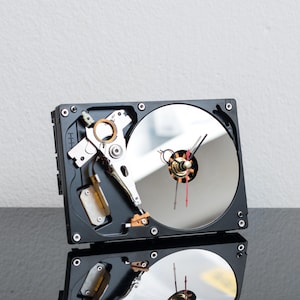 Könnte beinhalten: Eine Wanduhr aus einer wiederverwendeten Computerfestplatte. Das Gerät verfügt über eine kreisförmige, reflektierende silberne Scheibe als Zifferblatt, mit schwarzen Stunden- und Minutenzeigern sowie einem dünnen roten Sekundenzeiger.