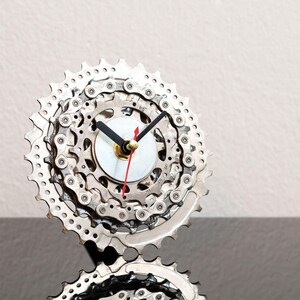 Può includere: Un orologio da parete circolare costruito con pignoni per bicicletta argentati impilati e un segmento di catena metallica. Il quadrante presenta un disco centrale bianco con due lancette nere e una sottile lancetta rossa dei secondi.