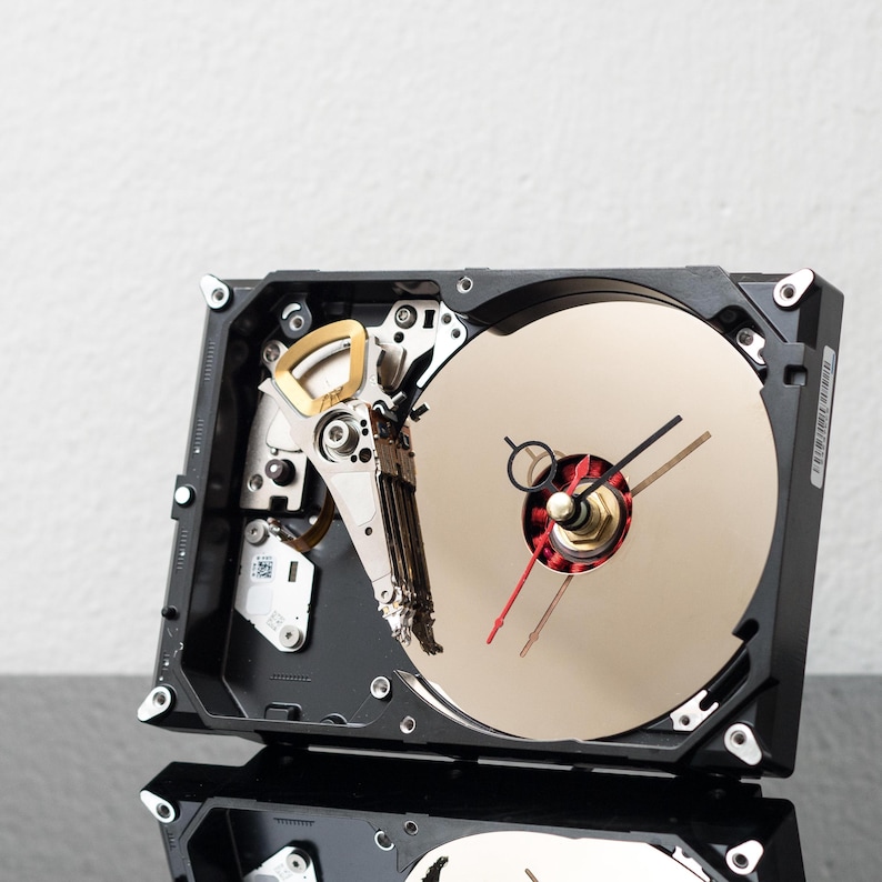 May include: A wall clock constructed from a repurposed black hard drive casing. The circular gold-toned platter serves as the clock face, featuring black and red clock hands. The internal mechanical arm remains visible in the upper left corner.