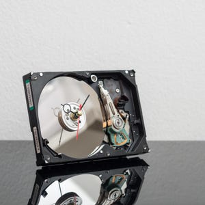Op de afbeelding: Een unieke klok gemaakt van een hergebruikte computer harde schijf. De wijzerplaat is een reflecterende schijf met wijzers en een klein, cirkelvormig klokmechanisme. De klok staat tegen een witte achtergrond.