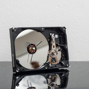 May include: A unique clock crafted from a repurposed computer hard drive. The clock face is the reflective surface of the drive, with clock hands indicating the time. The black frame and metallic components create an industrial aesthetic.