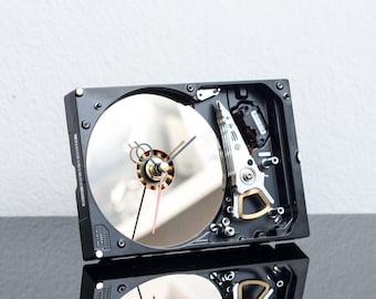 May include: A wall clock constructed from a repurposed black hard drive casing. The circular, reflective silver platter serves as the clock face, featuring black hour and minute hands along with a thin red second hand centered on the spindle.