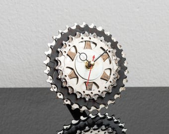 May include: A wall clock constructed from stacked silver and black metal bicycle gear sprockets. The clock face features a central silver disc with black hour and minute hands and a thin red second hand, all mounted on the circular gear assembly.