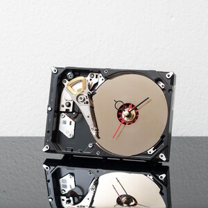 May include: A wall clock constructed from a repurposed hard drive, featuring a circular metallic platter as the clock face, a black and red clock hand mechanism, and exposed internal mechanical components within a rectangular black frame.