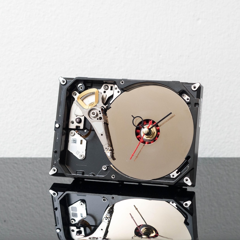 May include: A wall clock constructed from a repurposed hard drive, featuring a circular metallic platter as the clock face, a black and red clock hand mechanism, and exposed internal mechanical components within a rectangular black frame.