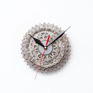 Può includere: Un'orologio da parete unico realizzato con ingranaggi di bicicletta riciclati. L'orologio presenta un design a ingranaggi argentati con lancette nere e una lancetta dei secondi rossa. L'orologio è montato su uno sfondo bianco.