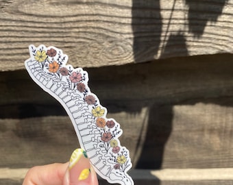 Op de afbeelding: Een sticker met een gestileerd wervelkolomontwerp met kleurrijke bloemen. De bloemen zijn in tinten geel, oranje en roze. De sticker wordt tegen een houten achtergrond gehouden.