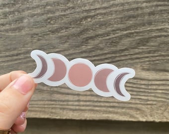 Puede incluir: Una pegatina con un diseño de cinco fases lunares en tonos rosa y marrón. Las fases lunares están dispuestas en forma de media luna.