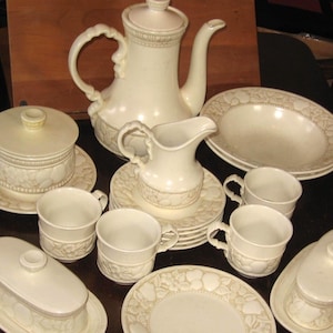 Könnte beinhalten: Ein cremefarbenes Vintage-Teeservice mit Teekanne, Sahnekännchen, Zuckerdose, Tassen, Untertassen und Servierschalen. Das Set hat ein geprägtes Blumendesign. Die Teekanne ist etwa 25 cm hoch.
