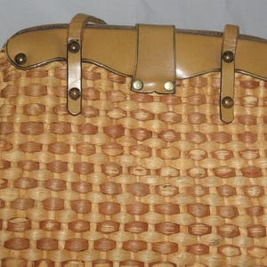 K&ouml;nnte beinhalten: Eine geflochtene Strohtasche mit einer braunen Ledertasche und Metallverzierungen. Die Tasche hat ein geflochtenes Muster und ein einzigartiges Design.