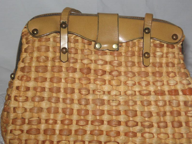 K&ouml;nnte beinhalten: Eine geflochtene Strohtasche mit einer braunen Ledertasche und Metallverzierungen. Die Tasche hat ein geflochtenes Muster und ein einzigartiges Design.