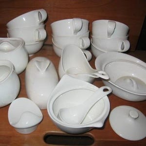 May include: A collection of white ceramic serving dishes and cups. The set includes several cups with handles, a sugar bowl, a creamer, a soup tureen with a ladle, and a covered dish. The items are arranged on a wooden surface.