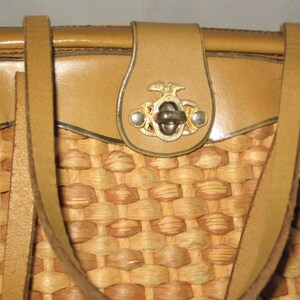 K&ouml;nnte beinhalten: Eine geflochtene Strohtasche mit braunen Lederriemen und einem dekorativen Metallverschluss. Die Tasche hat einen braunen Lederbesatz am oberen und unteren Rand.