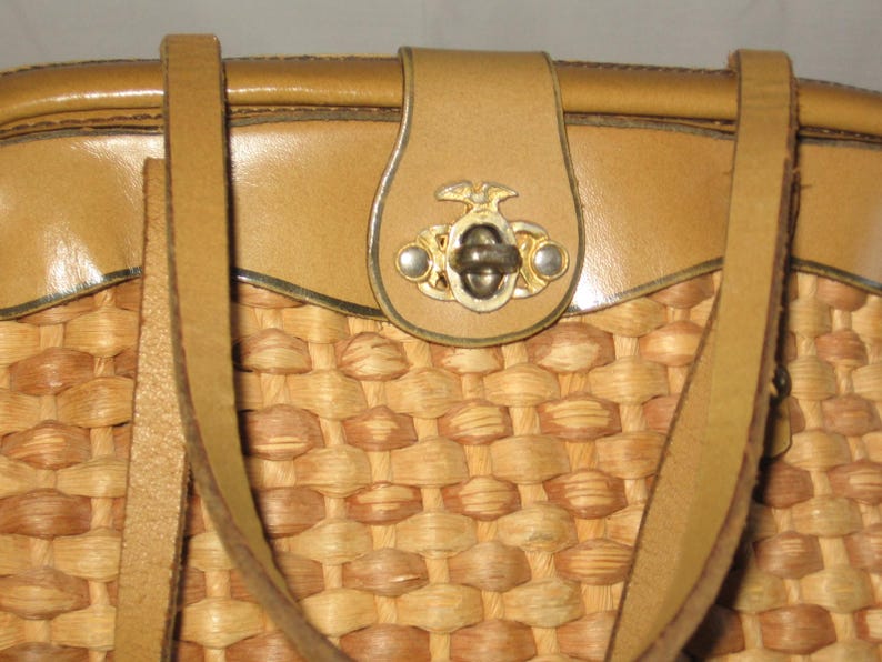 K&ouml;nnte beinhalten: Eine geflochtene Strohtasche mit braunen Lederriemen und einem dekorativen Metallverschluss. Die Tasche hat einen braunen Lederbesatz am oberen und unteren Rand.