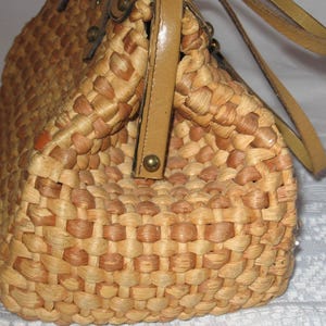 K&ouml;nnte beinhalten: Eine geflochtene Strohtasche mit einem braunen Lederriemen und goldenen Akzenten. Die Tasche ist aus nat&uuml;rlichen Materialien gefertigt und hat einen rustikalen, Vintage-Look.