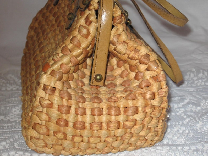 K&ouml;nnte beinhalten: Eine geflochtene Strohtasche mit einem braunen Lederriemen und goldenen Akzenten. Die Tasche ist aus nat&uuml;rlichen Materialien gefertigt und hat einen rustikalen, Vintage-Look.