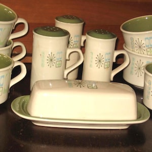 Puede incluir: Un juego de vajilla de cerámica crema vintage con un diseño moderno de mediados de siglo. El juego incluye tazas, saleros y pimenteros y una mantequera. Cada pieza presenta un interior y detalles en verde, con un patrón geométrico y de estrella estilizado en azul.