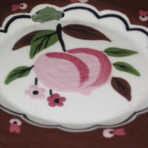 May include: A decorative plate with a scalloped edge and a brown rim. The center features a hand-painted peach design with pink and red accents, green leaves, and small pink flowers. The plate has a white interior.