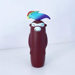 Könnte beinhalten: Eine marunfarbene, cartoonartige Figur eines Affen mit einem lächelnden Gesicht und einem regenbogenfarbenen Vogel obenauf. Die Affenfigur hat einen zylindrischen Körper mit Armen an den Seiten. Der Vogel ist blau, lila und gelb.