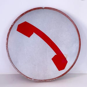 May include: A white circular metal sign with a red telephone icon. The sign has a red border and is slightly weathered.