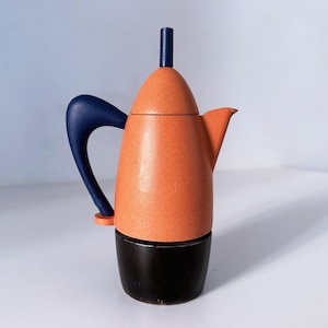 Pode incluir: Uma cafeteira com corpo superior e tampa em laranja texturizado, pega e botão da tampa em azul escuro e base preta lisa. A cafeteira apresenta uma forma cónica na tampa, uma pega curva e um pequeno bico vertedor no lado direito.