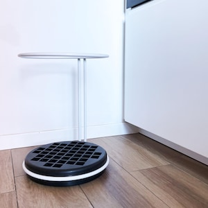 May include: A contemporary side table with a circular top and base. The top and the three supporting legs are light grey, while the base is black with a white trim. The base has a grid pattern. The table is on a wooden floor.