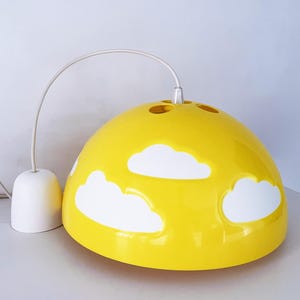 May include: A yellow pendant light with white cloud cutouts. The light has a rounded shape and a white cord and ceiling mount. The light is a cheerful addition to a child's room or nursery.