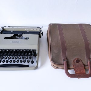 May include: A grey Olivetti Lettera 32 typewriter with a black keyboard and a brown leather carrying case. The typewriter has the text "Olivetti" and "Lettera 32" printed on the front.