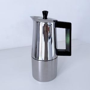 Pode incluir: Uma cafeteira italiana de aço inoxidável com alça e tampa pretas. A cafeteira tem uma superfície brilhante e reflexiva e uma base cilíndrica. A alça é retangular.